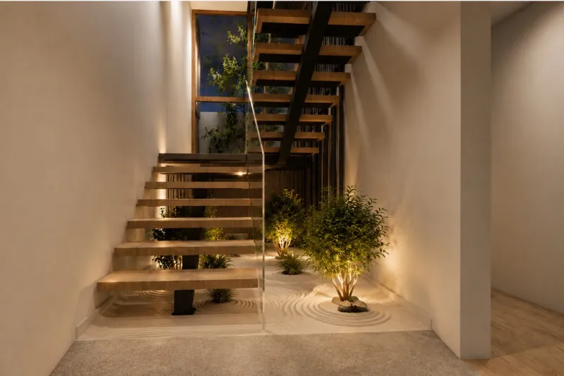 Escalera de peldaños volados sobre un jardín interior iluminado, con baranda de vidrio.